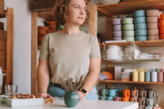 Serious concentrated curly businesswoman looking at camera, seller job, silicone tableware for kids. Workshop store, small business. Crafting design of pastel colorful bowls, plates, glasses, spoons