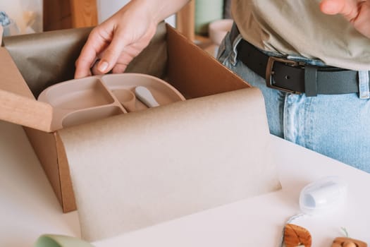 Cropped image of woman hands packing and wrapping shipment with silicone kids dishes in cardboard box for delivery. Wooden rattle toy, trinket decoration nearby on desk. Workshop store, work at home