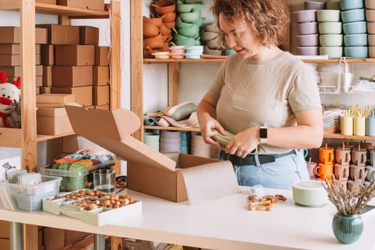 Smiling concentrated curly woman packing and wrapping shipment with silicone baby dish in cardboard box for delivery. Wooden rattle toy, trinket decoration nearby on desk. Workshop store, work at home