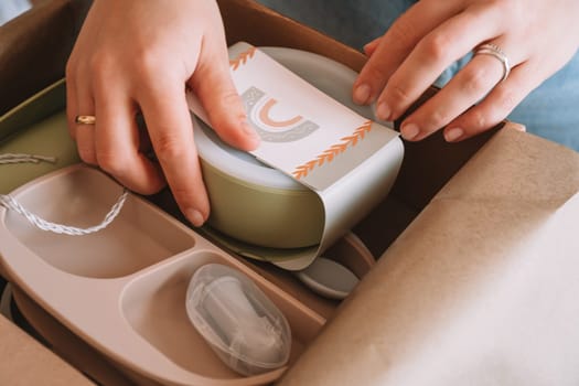 Cropped woman hands packing and wrapping shipment with silicone baby dish, plate, spoons, bowl, fixing her label to it, in cardboard box for delivery. copy space