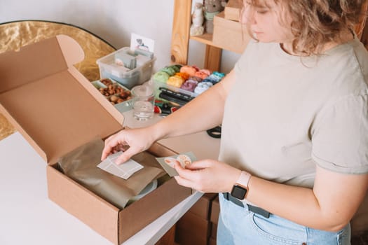 Smiling blond curly woman packing and wrapping shipment with postcards, silicone baby dishes in cardboard delivery box. Colorful skein of thread in container on desk. Workshop store, small business