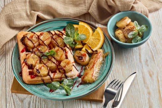 Delicious waffles with caramelized banana, sliced orange, mint, jam and sweet sauce on plate on light wooden background. Top view. Table setting. Dessert. Serving food
