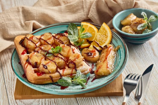 Delicious waffles with caramelized banana, sliced orange, mint, jam and sweet sauce on plate on light wooden background. Table setting. Dessert. Serving food