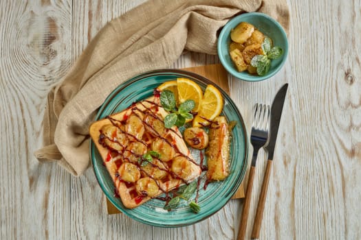 Delicious waffles with caramelized banana, sliced orange, mint, jam and sweet sauce on plate on light wooden background. Top view. Table setting. Dessert. Serving food