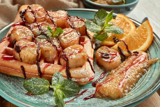 Delicious waffles with caramelized banana, sliced orange, mint, jam and sweet sauce on plate on light wooden background. Table setting. Dessert. Serving food