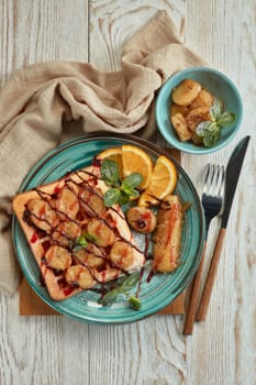 Delicious waffles with caramelized banana, sliced orange, mint, jam and sweet sauce on plate on light wooden background. Top view. Table setting. Dessert. Serving food