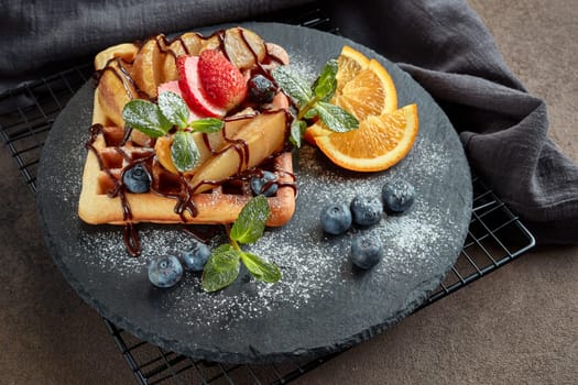 Delicious waffles with caramelized pear, berries, mint, powdered sugar and sweet sauce on black serving board on dark brown background. Dessert. Serving food. Top view