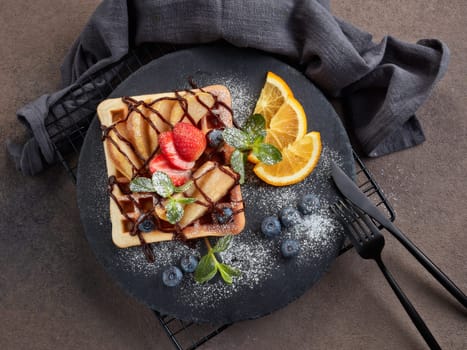 Delicious waffles with caramelized pear, berries, mint, powdered sugar and sweet sauce on black serving board on dark brown background. Dessert. Serving food. Top view