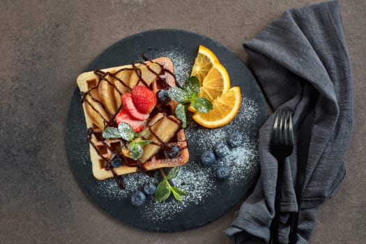 Delicious waffles with caramelized pear, berries, mint, powdered sugar and sweet sauce on black serving board on dark brown background. Dessert. Serving food. Top view