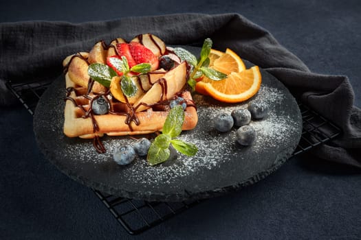 Delicious waffles with caramelized pear, berries, mint, powdered sugar and sweet sauce on black serving board on dark brown background. Dessert. Serving food. Table setting