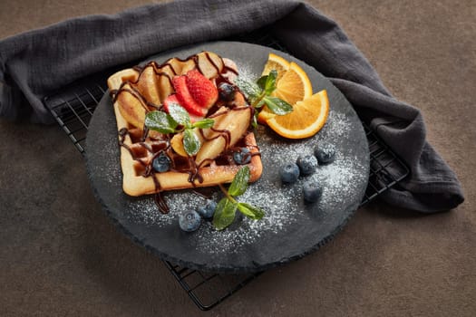 Delicious waffles with caramelized pear, berries, mint, powdered sugar and sweet sauce on black serving board on dark brown background. Dessert. Serving food. Top view
