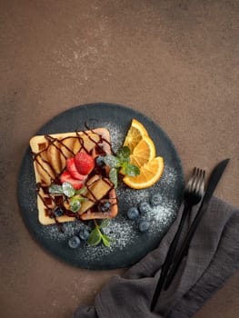 Delicious waffles with caramelized pear, berries, mint, powdered sugar and sweet sauce on black serving board on dark brown background. Dessert. Serving food. Top view
