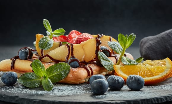 Delicious waffles with caramelized pear, berries, mint, powdered sugar and sweet sauce on black serving board on grey background. Dessert. Serving food. Table setting