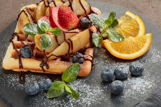 Delicious waffles with caramelized pear, berries, mint, powdered sugar and sweet sauce on black serving board on brown background. Dessert. Serving food. Table setting