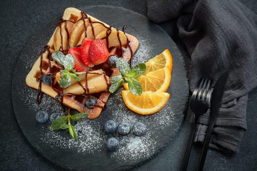 Delicious waffles with caramelized pear, berries, mint, powdered sugar and sweet sauce on black serving board on grey background. Dessert. Serving food. Table setting