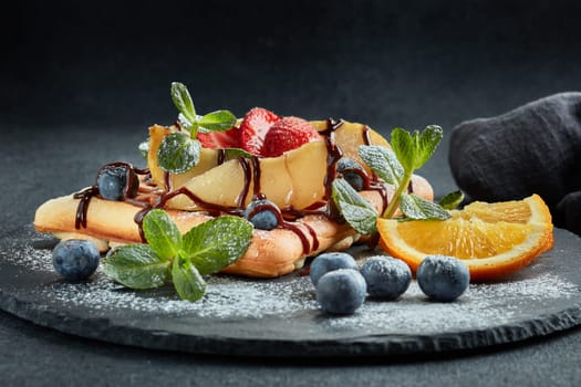 Delicious waffles with caramelized pear, berries, mint, powdered sugar and sweet sauce on black serving board on grey background. Dessert. Serving food. Table setting