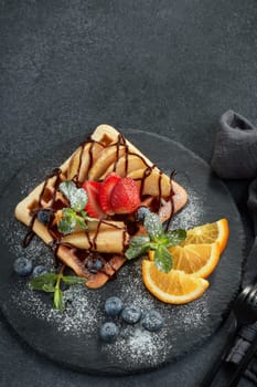 Delicious waffles with caramelized pear, berries, mint, powdered sugar and sweet sauce on black serving board on grey background. Dessert. Serving food. Table setting