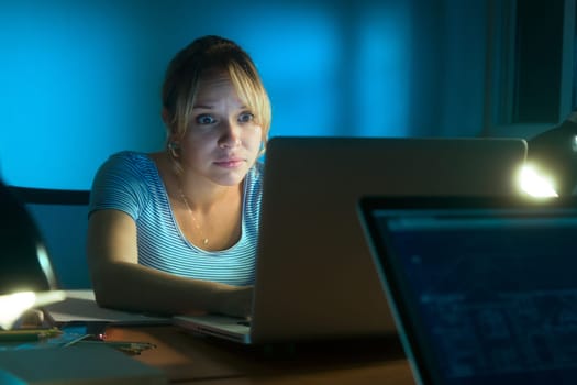 Beautiful woman working late at night in office, surfing the web and reading a post on social network with laptop computer. The girl looks at the screen with scared expression