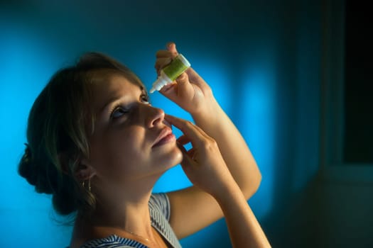 Beautiful young latina woman with eyesight problems, working late at night in office. The girl puts collyrium drops in tired eyes and smiles.
