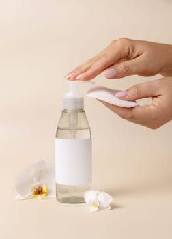 A woman hand pumping liquid on a make-up remover pad from a cosmetic bottle near orchid flowers on beige close up, label mockup. Skincare beauty product package, minimal composition..