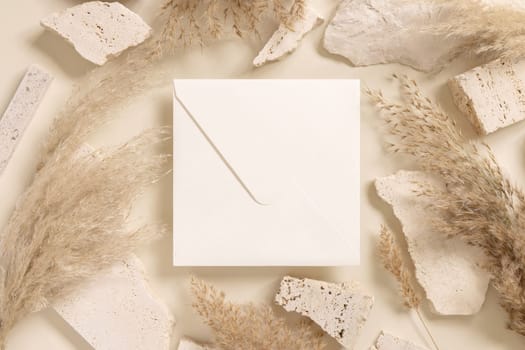 Blank square envelope near beige travertine stones and boho dried pampas grass top view, mockup. Bohemian greeting or wedding Invitation. Close to nature.