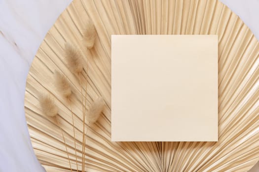 Blank card on dry beige palm leaf near hare's tail grass top view, mockup. Boho scene with square paper card. Bohemian or close to nature greeting or wedding Invitation. Minimal composition.
