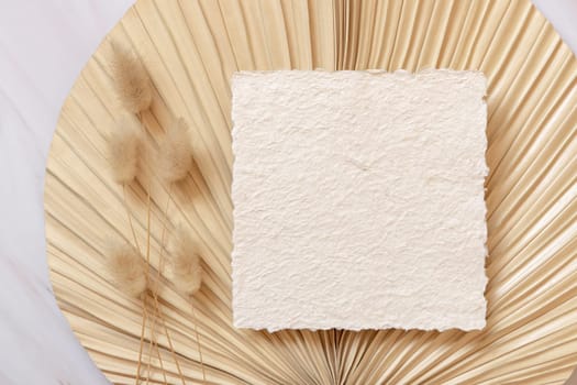 Blank handmade paper card on dry beige palm leaf near hare's tail grass top view, mockup. Boho scene with square card. Bohemian or close to nature greeting card or wedding Invitation.