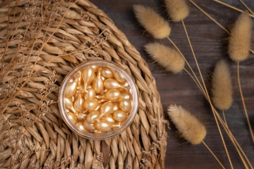 Beauty serum capsules in a jar on wattled boho placemat on wood near Hare's Tail grass top view. Natural skincare product for everyday beauty routine. Bohemian eco friendly cosmetics flat lay.