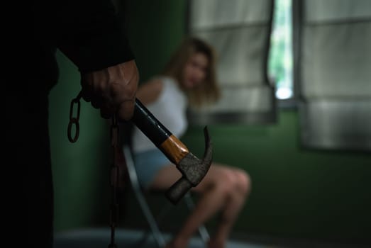 Man hand holding hammer and woman sitting on chair and tied with ropes in dark room
