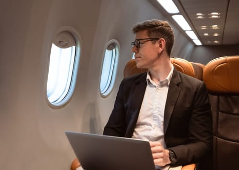 Businessman in formal wear and glasses looking out of aircraft window during flight while working with laptop computer. Internet connection on board service and business travel concept