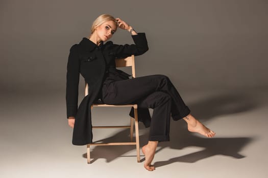 Beauty blond woman  in a black coat on chair on a gray background
