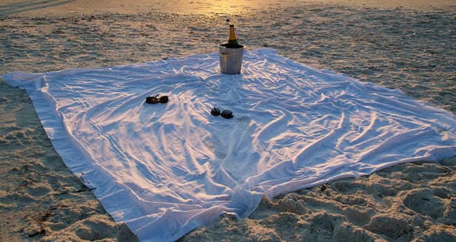 A big white sheet is laid out on the beach and there is an ice bucket and bottle of champagne with two pairs of sunglasses at sunset.
