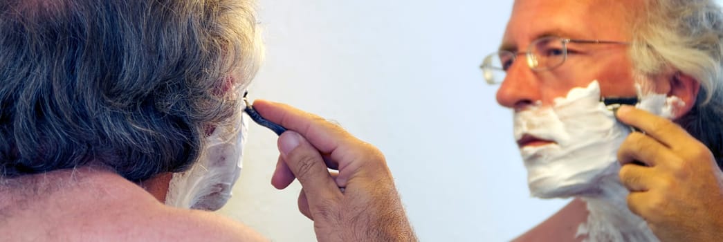 A man with eyeglasses has shaving cream on his face and looking in the mirror is beginning to shave.