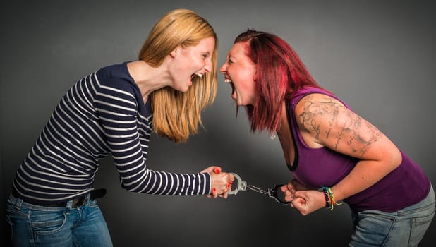 Two angry women screaming und fighting with a pair of handcuffs  on grey background
