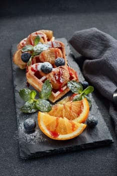 Delicious waffles with caramelized pear, berries, mint, orange and sweet sauce on black serving board on grey background. Top view. Sweet meal. Dessert. Serving food. Table setting