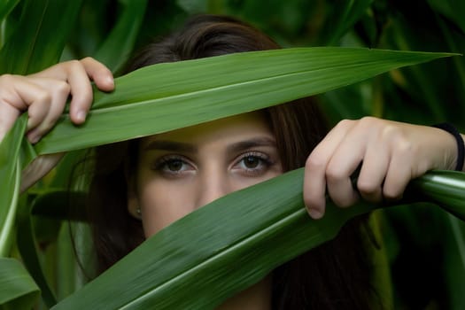Portrait of beautiful young girl hiding by green leaves. Horizontally.