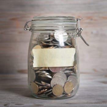 Coins in glass money jar with blank label, financial concept. Vintage wooden background with dramatic light.