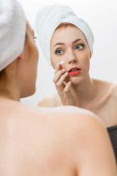Young woman with towel in her hair carries cream on her face on in the mirror