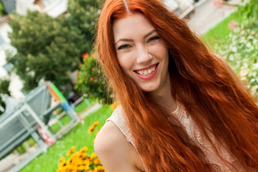 attractive young woman with long red hair sits in the garden on a wall and relaxes in summer in daylight and good weather