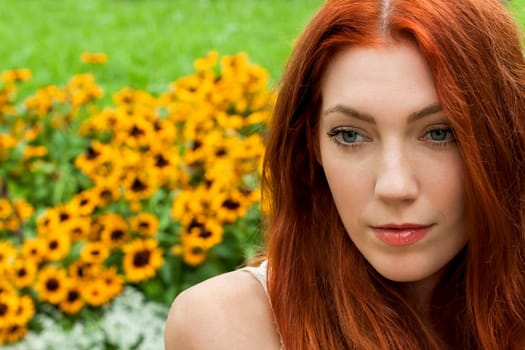 attractive young woman with long red hair sits in the garden on a wall and relaxes in summer in daylight and good weather