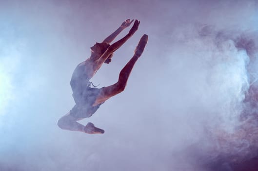 young ballet dancer jumping on a lilac background. Ballerina is wearing in blue dress and pointe shoes. The outline shooting - silhouette of girl with smoke effect