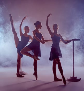 Three young ballerinas  in blue dresses stretching on the bar on lilac background
