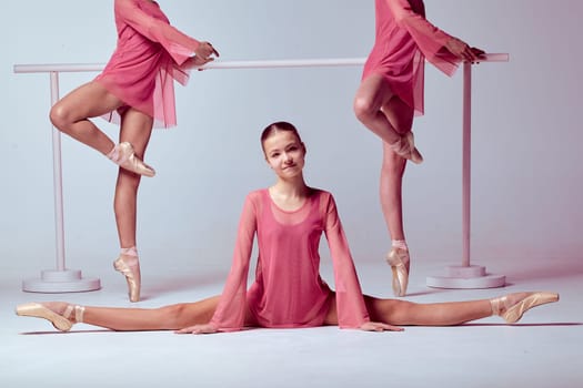 Three young ballerinas  in a pink dresses stretching on the bar on beige background