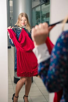Woman trying red dress shopping for clothing. Beautiful happy blonde smiling young shopper woman looking in mirror standing in clothes store.