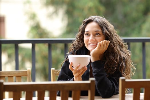 Happy woman holding bowl contemplating at breakfast in a balcony