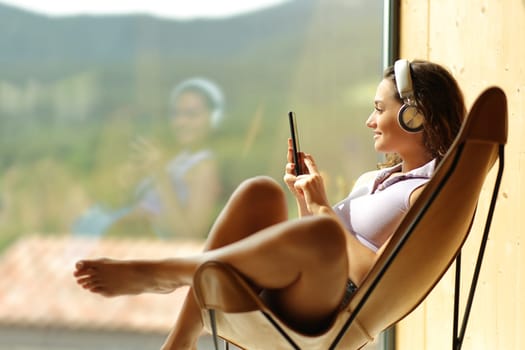 Happy woman at home or hotel listening to music sitting on a chair looking through a window
