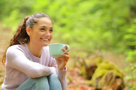 Happy woman sitting in a forest drinking coffee looking away