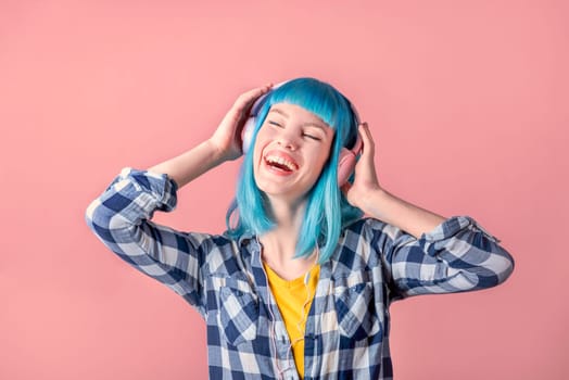 Beautiful smiling young woman with blue color dyed hair with pink headphones listening to music and dance in front of pastel studio background.