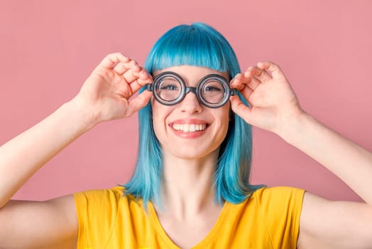 Beautiful young woman with blue color dyed hair and hipster funny goggles smiling in front of pastel studio background.