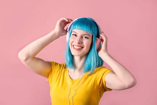 Beautiful smiling young woman with blue color dyed hair with pink headphones listening to music and dance in front of pastel studio background.